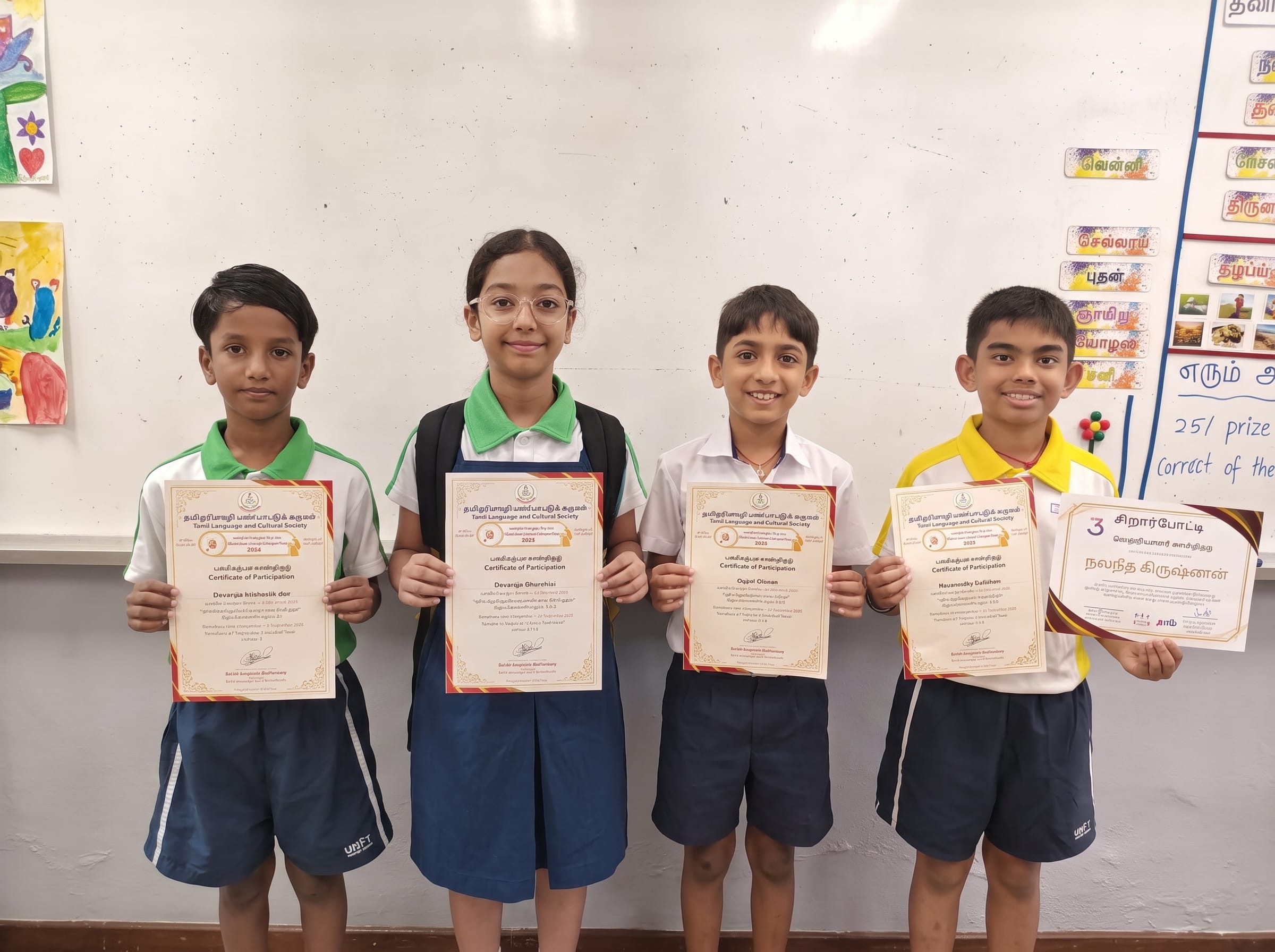 A group photo of four smiling students standing side-by-side in a classroom, proudly holding their Tamil literary competition certificates in front of a whiteboard with colorful student artwork.